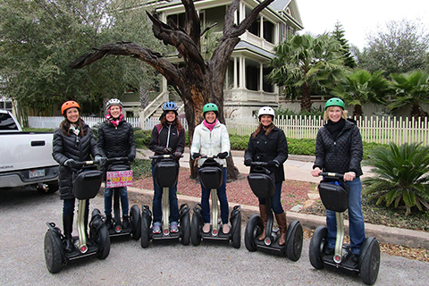 A group of people pose in front of a carved tree while on the Tree Carvings Segway Tour from SegCity in Galveston, Texas, which is available for free to guests staying at Xplorie participating properties.